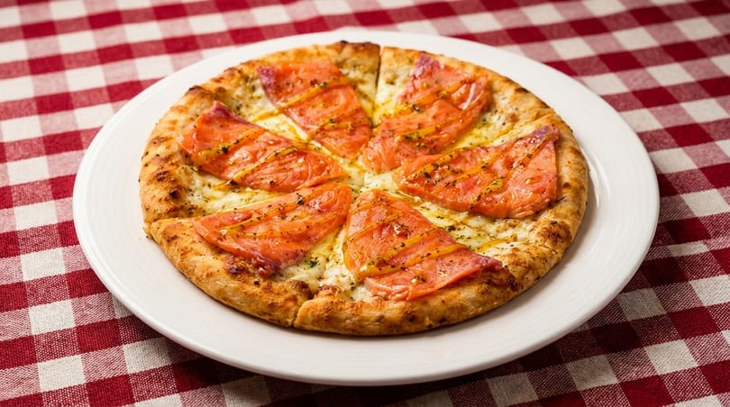 A salmon pizza with cheese and herbs on a white plate, resting on a red and white checkered tablecloth.