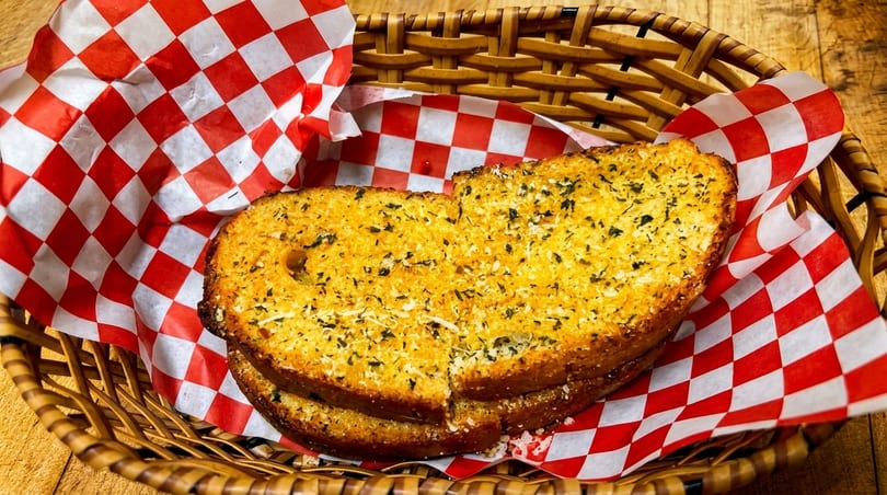 Golden-brown garlic bread slices with herbs and cheese in a wicker basket lined with red and white checkered paper.