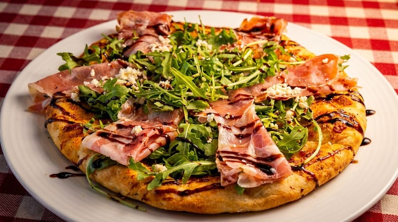 A flatbread pizza topped with prosciutto, arugula, crumbled cheese, and balsamic glaze on a red and white checkered tablecloth.
