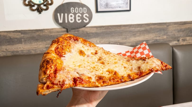Hand holding a large slice of cheese pizza on a white plate with a checkered paper liner.